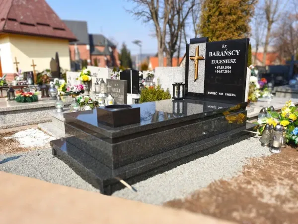 Masywny nagrobek z czarnego granitu ze złotym krzyżem na jasnej, groszkowanej wstawce i grawerowanymi napisami